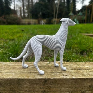 May include: A gray, stylized sculpture of a greyhound dog. The dog is standing on a wooden surface, with a green grassy background. The sculpture has a unique, layered design, with horizontal lines creating a textured effect.