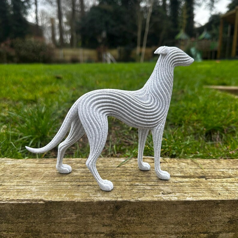 May include: A gray, stylized sculpture of a greyhound dog. The dog is standing on a wooden surface, with a green grassy background. The sculpture has a unique, layered design, with horizontal lines creating a textured effect.