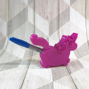 Puede incluir: Un portalápices de unicornio impreso en 3D de color rosa con un bolígrafo azul que sobresale de la parte posterior. El unicornio está mirando hacia el lado derecho de la imagen.