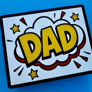 May include: A rectangular sign with the word "DAD" in large yellow letters, outlined in black, inside a white speech bubble with a red outline. The sign has a black border and yellow stars on a blue background.
