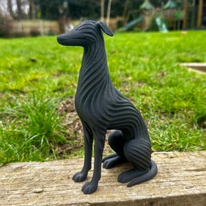 May include: A black, stylized sculpture of a dog in a seated position. The dog has a long neck and a slender body with a textured, wave-like pattern. The sculpture is placed on a wooden surface with a green grassy background.