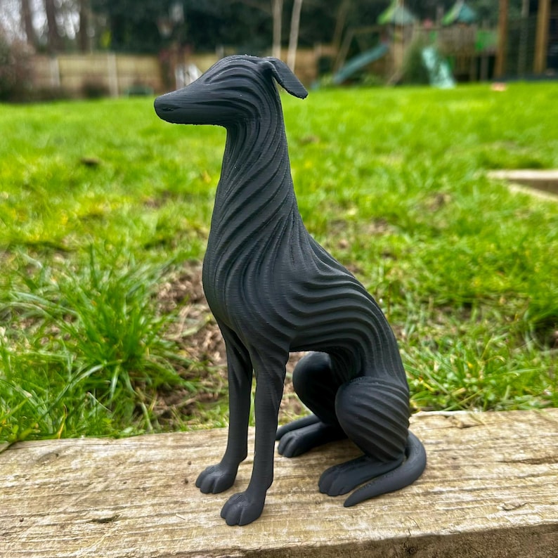 May include: A black, stylized sculpture of a dog in a seated position. The dog has a long neck and a slender body with a textured, wave-like pattern. The sculpture is placed on a wooden surface with a green grassy background.