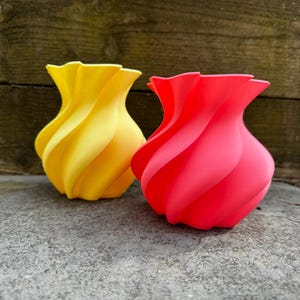May include: Two decorative vases with a spiral design. One vase is bright yellow, and the other is a vibrant red. The vases have a smooth, matte finish and are placed on a gray surface. The vases are suitable for home decor.