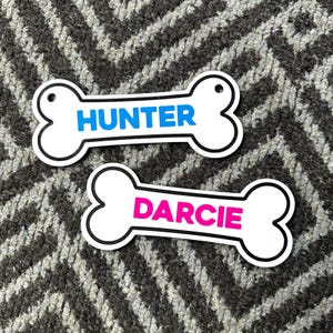 Puede incluir: Dos etiquetas blancas para mascotas con forma de hueso de perro y borde negro. Una etiqueta tiene el nombre "HUNTER" en azul y la otra "DARCIE" en rosa. Ambas etiquetas tienen un agujero para sujetarlas al collar.