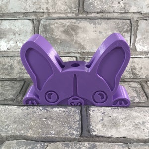 May include: A purple 3D printed figurine of a French bulldog peeking over a brick wall. The figurine has a playful expression with large ears and a cute nose.