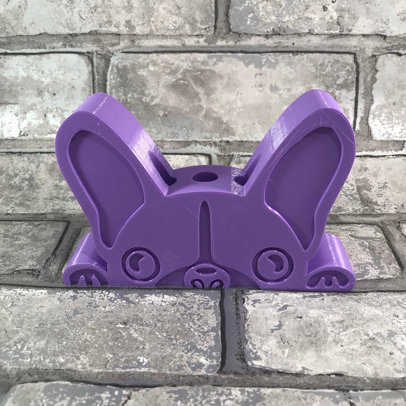 May include: A purple 3D printed figurine of a French bulldog peeking over a brick wall. The figurine has a playful expression with large ears and a cute nose.