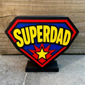 May include: A black, diamond-shaped sign with the word "SUPERDAD" in yellow, outlined in black, set against a blue background. The sign has a red border and a yellow starburst design. It stands on a small black base.