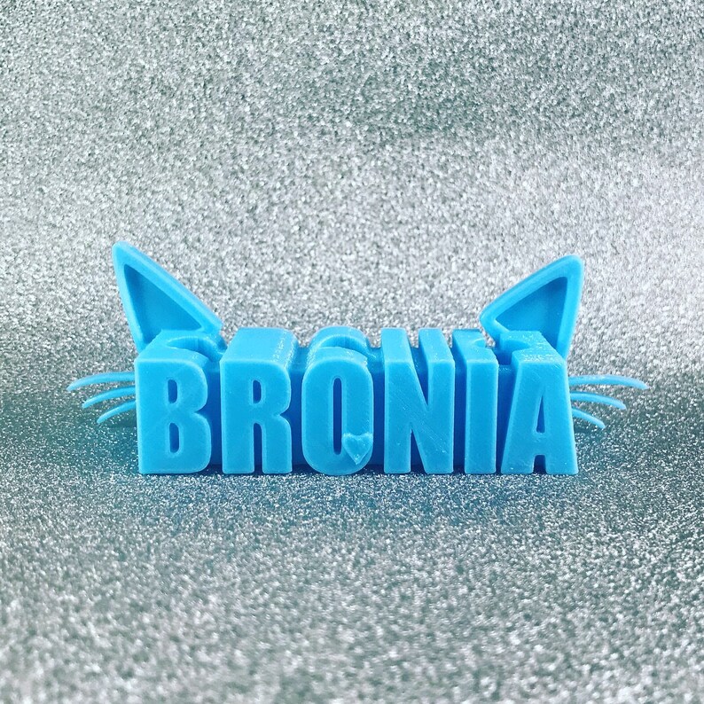 May include: A light blue 3D printed cat figurine with the name "BRONIA" spelled out in block letters.