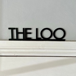Könnte beinhalten: Schwarzes Holzschild mit dem Text "THE LOO" in einer modernen Schriftart.