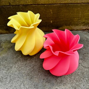 May include: Two decorative vases with a unique, swirling design. One vase is bright yellow, and the other is a vibrant pink. The vases have a textured, petal-like appearance, adding a modern touch to any space.