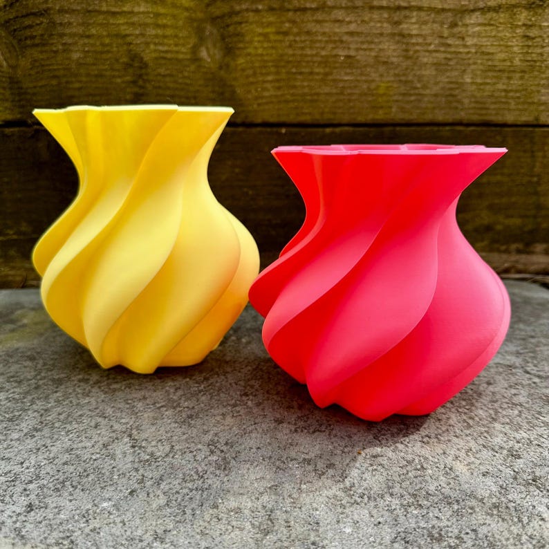May include: Two decorative vases with a unique, swirling design. One vase is bright yellow, and the other is a vibrant red. The vases are made of a smooth material and sit on a gray surface. The vases have a modern, artistic look.