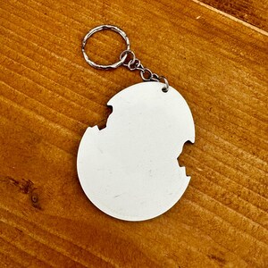 Może przedstawiać: Biały, okrągły brelok do kluczy ze srebrnym k&oacute;łkiem i łańcuszkiem. Brelok ma unikalny, nieregularny kształt z brakującym fragmentem na krawędzi. Tło to drewniana powierzchnia.