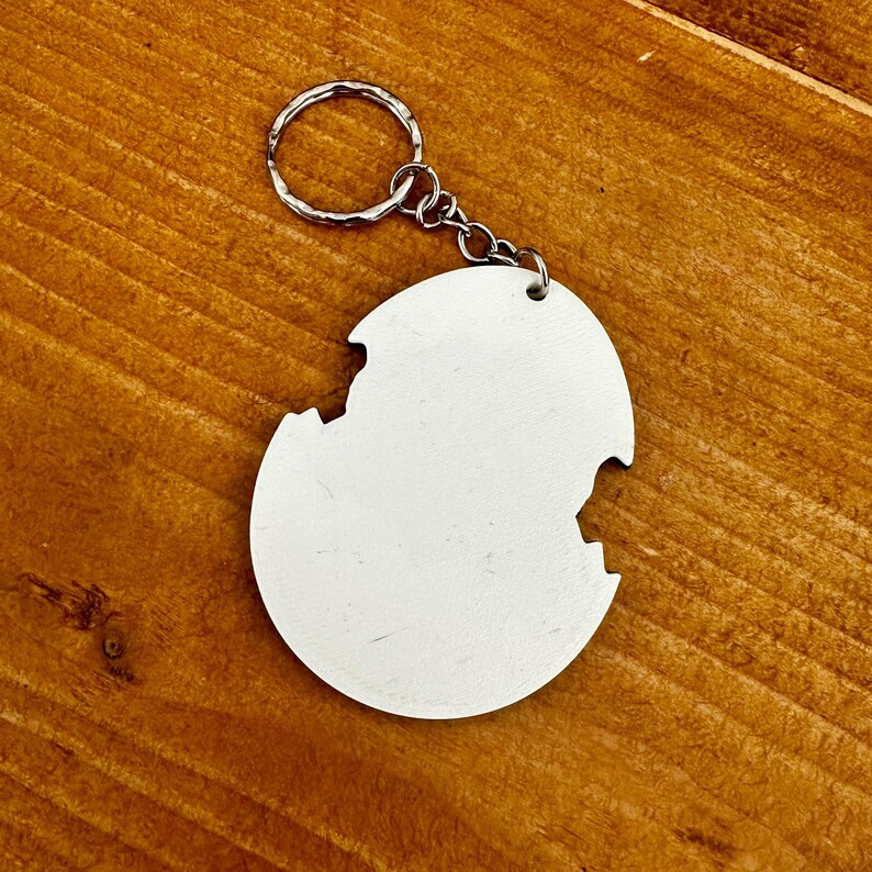 Może przedstawiać: Biały, okrągły brelok do kluczy ze srebrnym k&oacute;łkiem i łańcuszkiem. Brelok ma unikalny, nieregularny kształt z brakującym fragmentem na krawędzi. Tło to drewniana powierzchnia.