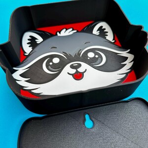 May include: A black and red container with a raccoon face design. The raccoon has a gray face, white accents, and a red tongue. The container has a matching lid with a blue accent.
