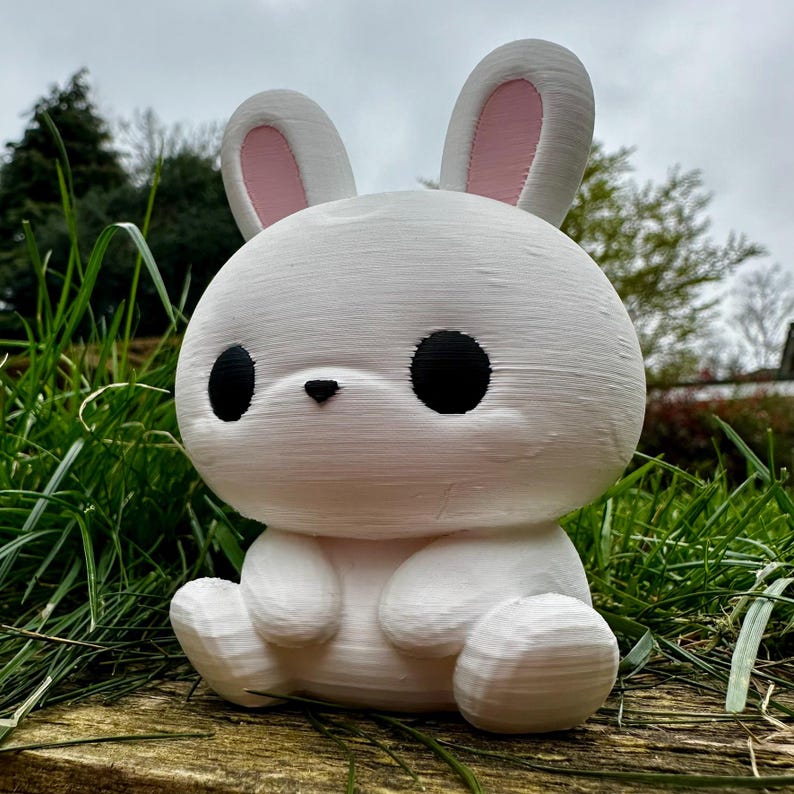 Puede incluir: Una figura de conejo blanco con grandes ojos negros y orejas internas rosas. El conejo est&aacute; sentado sobre una superficie de madera, rodeado de hierba verde. La figura tiene un dise&ntilde;o lindo y de dibujos animados.