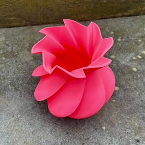May include: A vibrant pink, flower-shaped decorative vase. The vase has a layered petal design, with a wide opening at the top. The vase is made of a smooth material and sits on a gray surface.