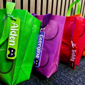 Puede incluir: Primer plano de tres bolsas de regalo coloridas: verde, morado y rojo. Cada bolsa tiene un asa a juego y una etiqueta personalizada con un nombre y un icono de mando de videojuegos. Las bolsas tienen un patr&oacute;n circular.