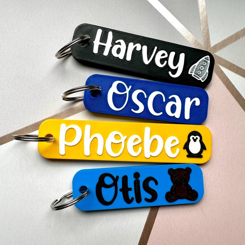 Puede incluir: Cuatro llaveros rectangulares en negro, azul, amarillo y azul. Cada llavero tiene un nombre en blanco, con un peque&ntilde;o gr&aacute;fico. Los nombres son Harvey, Oscar, Phoebe y Otis. Harvey tiene un cohete, Phoebe tiene un ping&uuml;ino y Otis tiene un oso de peluche.