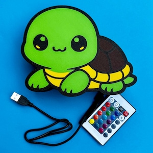 K&ouml;nnte beinhalten: Gr&uuml;ne, schildkr&ouml;tenf&ouml;rmige LED-Lampe mit braunem Panzer und gelben Akzenten. Inklusive USB-Kabel und Fernbedienung mit verschiedenen Farboptionen. Die Lampe befindet sich auf blauem Hintergrund.