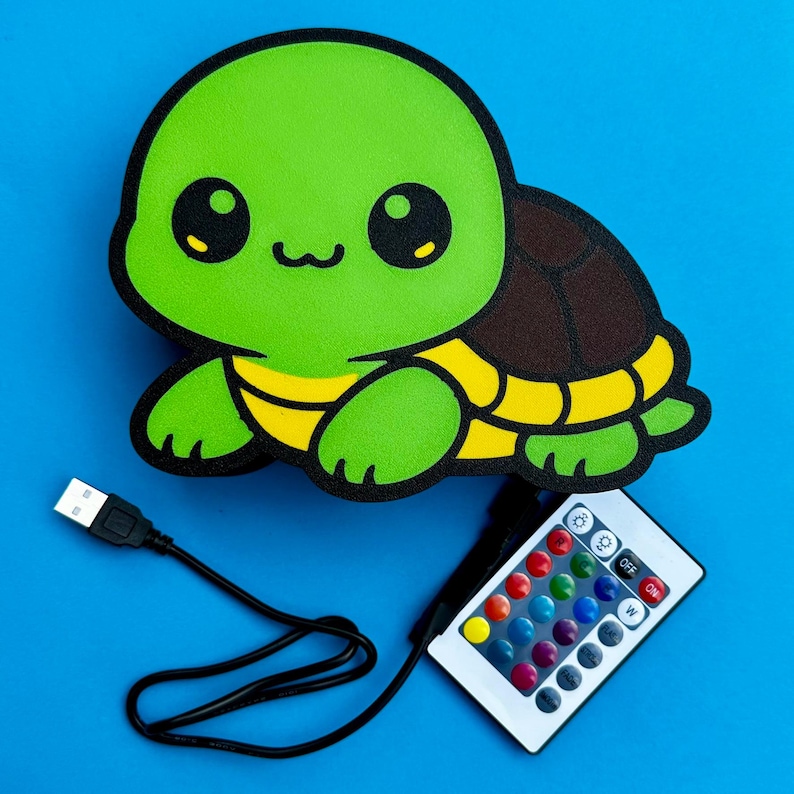 K&ouml;nnte beinhalten: Gr&uuml;ne, schildkr&ouml;tenf&ouml;rmige LED-Lampe mit braunem Panzer und gelben Akzenten. Inklusive USB-Kabel und Fernbedienung mit verschiedenen Farboptionen. Die Lampe befindet sich auf blauem Hintergrund.