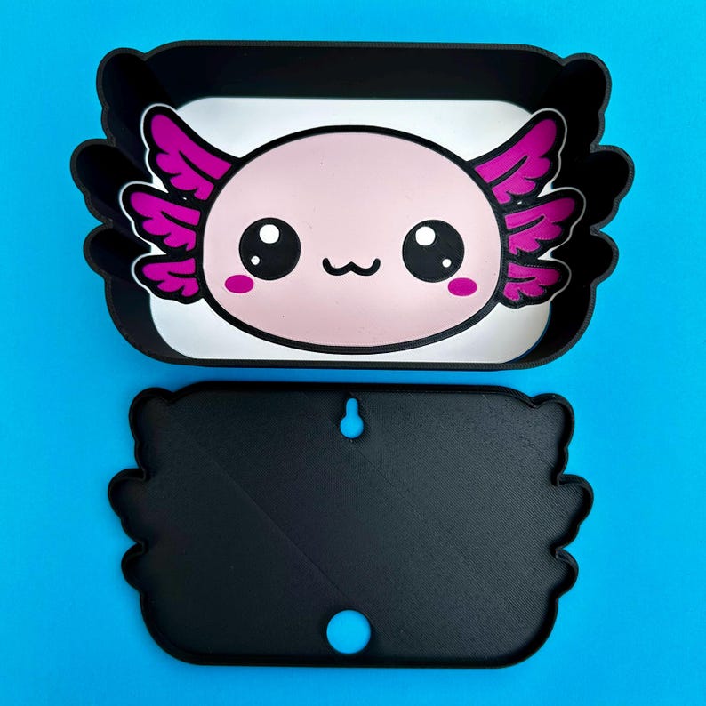 K&ouml;nnte beinhalten: Ein schwarz-wei&szlig;es Tablett im Axolotl-Design und eine passende Wandhalterung. Das Tablett zeigt ein rosa Axolotl-Gesicht mit gro&szlig;en schwarzen Augen, rosa Wangen und lila fl&uuml;gelartigen Kiemen. Die Wandhalterung ist schwarz mit einem Loch zum Aufh&auml;ngen.