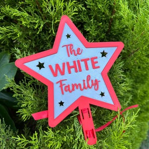 Puede incluir: Una decoraci&oacute;n en forma de estrella roja y azul claro con el texto "The WHITE Family". La estrella tiene forma de estrella recortada y un clip rojo. La decoraci&oacute;n se muestra sobre un fondo de follaje verde.