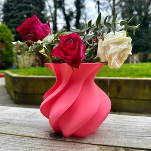 May include: A vibrant pink vase with a unique, swirling design holds a bouquet of fresh flowers. The arrangement includes deep red roses, a creamy white rose, and green foliage. The vase sits on a weathered wooden surface.