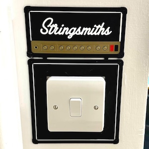 Op de afbeelding: Een zwart-witte lichtschakelaar-afdekplaat met de woorden "Stringsmithes" erop gedrukt. De afdekplaat is gevormd als een gitaarversterker met een rij knoppen.