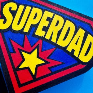 May include: A rectangular patch with the words "SUPERDAD" in large yellow letters. The patch has a blue background with a red and yellow starburst design and a yellow star in the center. The border is red and black.