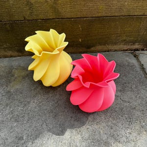 May include: Two decorative vases, one yellow and one pink, sit side-by-side on a gray stone surface. The vases have a unique, petal-like design, with a ruffled top. The yellow vase is on the left, and the pink vase is on the right.