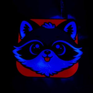 May include: A raccoon face design with blue neon highlights on a red square background. The raccoon has large eyes, a small pink tongue, and a black and blue color scheme. The image is set against a black background.