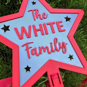 Puede incluir: Una decoraci&oacute;n en forma de estrella roja y azul claro con el texto "The WHITE Family". La estrella tiene formas de estrella recortadas y una cinta roja para colgar. La decoraci&oacute;n es probablemente para una fiesta u ocasi&oacute;n especial.