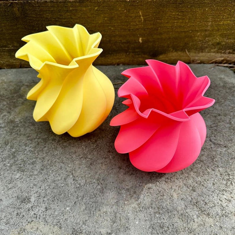 May include: Two decorative vases. One is yellow, the other is pink. Both have a unique, petal-like design around the top and a rounded base. The vases are made of a smooth material and sit on a gray surface.