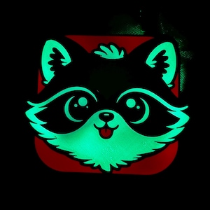 May include: A glowing raccoon face design with a red background. The raccoon has large, round eyes, a small nose, and a smiling mouth. The outline and details of the raccoon glow green against the black background.