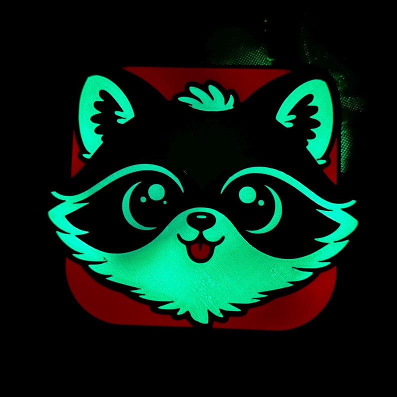 May include: A glowing raccoon face design with a red background. The raccoon has large, round eyes, a small nose, and a smiling mouth. The outline and details of the raccoon glow green against the black background.