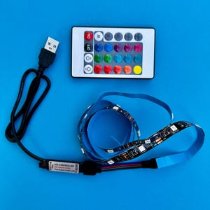 May include: LED strip lights with a remote control and USB cable, displayed on a blue surface. The remote is white with colored buttons and an "ON" and "OFF" button. The LED strip is black with small lights and a blue backing.