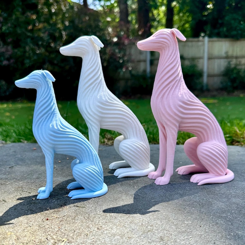 May include: Three decorative greyhound dog sculptures in pastel colors: light blue, white, and pink. Each dog is in a seated position with a textured, ridged design. The sculptures are displayed outdoors on a concrete surface.