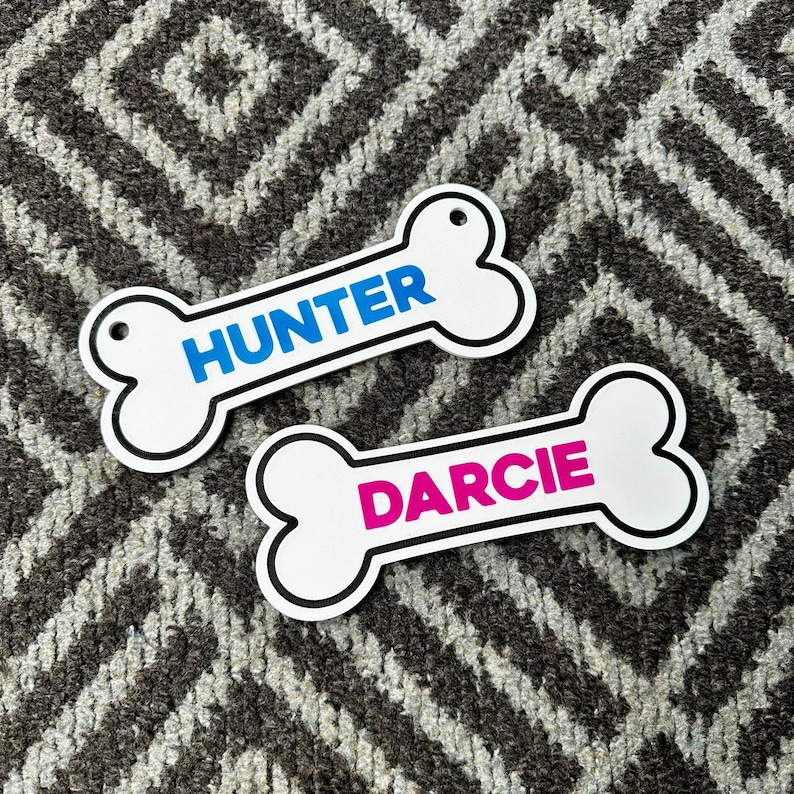 Puede incluir: Dos etiquetas blancas en forma de hueso para mascotas con contornos negros. Una etiqueta tiene "HUNTER" en azul, la otra "DARCIE" en rosa. Ambas etiquetas tienen un agujero para sujetar.