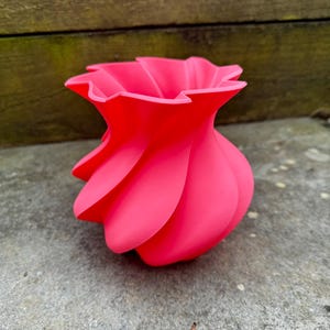May include: A vibrant, hot pink vase with a unique, swirling design. The vase has a ruffled top and a smooth, matte finish. The spiraling shape creates an eye-catching, modern aesthetic. This decorative piece is suitable for displaying flowers or as a standalone art object.