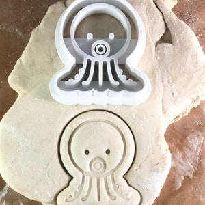 May include: White plastic cookie cutter shaped like an octopus with a round head and eight tentacles. The cutter is on top of a piece of white dough.