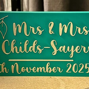 Op de afbeelding: Een turquoise rechthoekig bord met gouden letters met de tekst "Mrs & Mrs Childs-Sayers 4th November 2025". Een gouden bladontwerp staat in de linkerbovenhoek. Het bord is waarschijnlijk voor een bruiloft.