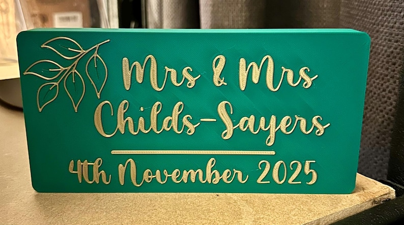 Op de afbeelding: Een turquoise rechthoekig bord met gouden letters met de tekst "Mrs & Mrs Childs-Sayers 4th November 2025". Een gouden bladontwerp staat in de linkerbovenhoek. Het bord is waarschijnlijk voor een bruiloft.