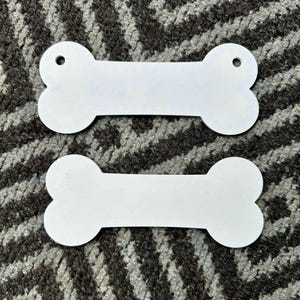 Puede incluir: Dos etiquetas blancas en forma de hueso con un agujero en un extremo. Las etiquetas est&aacute;n hechas de un material liso y s&oacute;lido y est&aacute;n colocadas sobre un fondo estampado. Estas etiquetas son adecuadas para productos para mascotas.