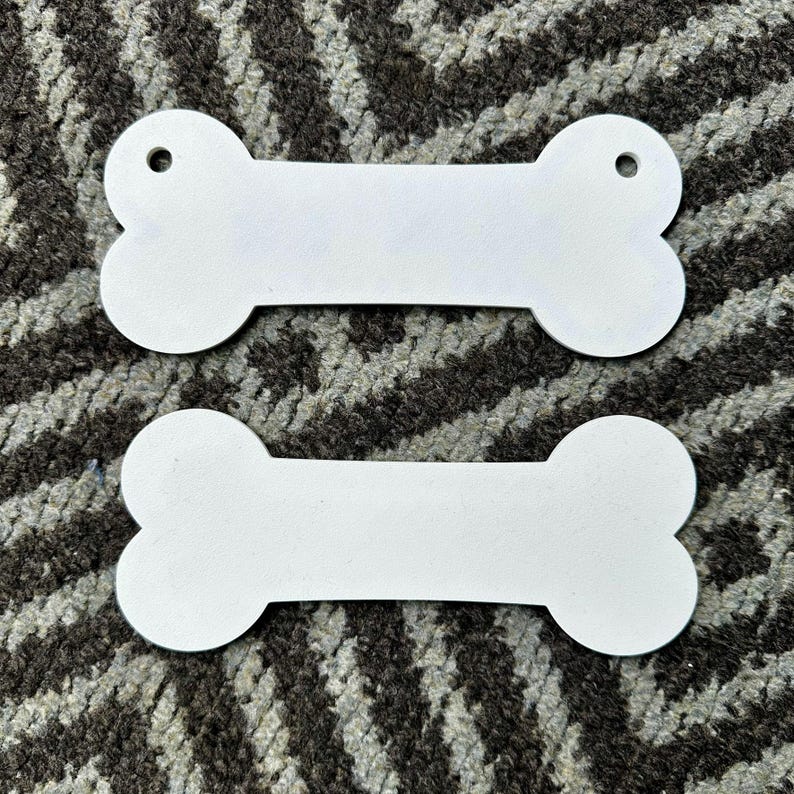 Puede incluir: Dos etiquetas blancas en forma de hueso con un agujero en un extremo. Las etiquetas est&aacute;n hechas de un material liso y s&oacute;lido y est&aacute;n colocadas sobre un fondo estampado. Estas etiquetas son adecuadas para productos para mascotas.