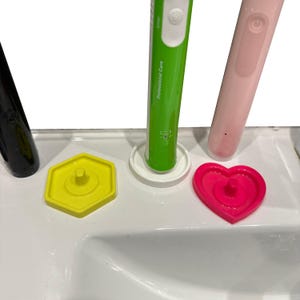 May include: A close-up of a white sink with three electric toothbrushes and three colored toothbrush head covers. The toothbrushes are green, pink, and black. The covers are yellow, white, and pink.