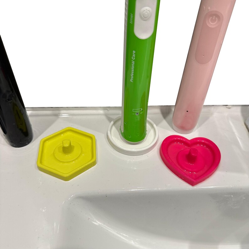 May include: A close-up of a white sink with three electric toothbrushes and three colored toothbrush head covers. The toothbrushes are green, pink, and black. The covers are yellow, white, and pink.