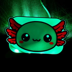 K&ouml;nnte beinhalten: Ein leuchtendes gr&uuml;nes Nachtlicht in Form eines Axolotl. Der Cartoon-Axolotl hat gro&szlig;e schwarze Augen, rote federartige Kiemen und einen l&auml;chelnden Mund. Das Licht ist rechteckig mit abgerundeten Ecken und einem schwarzen Umriss. Der Hintergrund ist dunkel.