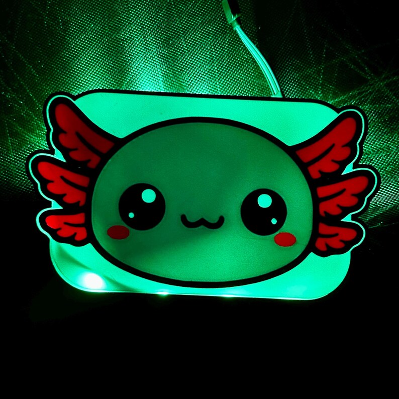 K&ouml;nnte beinhalten: Ein leuchtendes gr&uuml;nes Nachtlicht in Form eines Axolotl. Der Cartoon-Axolotl hat gro&szlig;e schwarze Augen, rote federartige Kiemen und einen l&auml;chelnden Mund. Das Licht ist rechteckig mit abgerundeten Ecken und einem schwarzen Umriss. Der Hintergrund ist dunkel.