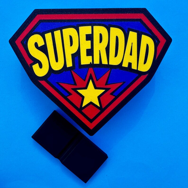 May include: A superhero-themed emblem with the text "SUPERDAD" in yellow, set against a blue background within a red-bordered diamond shape. A yellow starburst is in the center. A black rectangular object is below the emblem.