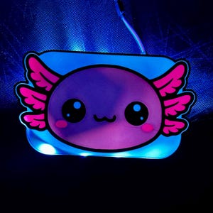 K&ouml;nnte beinhalten: Ein leuchtendes LED-Licht in Form eines Axolotls. Der Axolotl hat einen lila K&ouml;rper, rosa Kiemen und gro&szlig;e schwarze Augen. Es ist auf einer blauen rechteckigen Basis mit blau leuchtender Umrandung angebracht. Ein d&uuml;nner Draht erstreckt sich von oben.